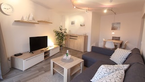 A modern living room with a flat-screen TV, a wooden coffee table, a grey sofa, and a small desk with a chair. - Villa am Meer App. 5 incl. WLAN (Grömitz)
