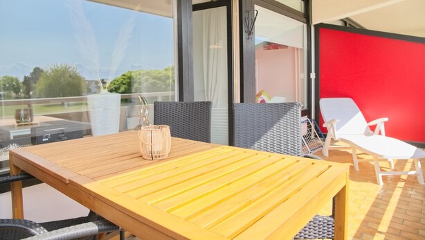 Un balcón con una mesa de madera, sillas de mimbre y un separador rojo.