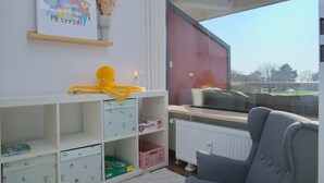 Una habitación con un ventanal grande, un sofá gris, una estantería blanca con juguetes infantiles y un cuadro en la pared.