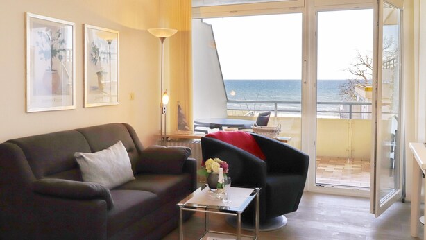 Un salon moderne avec un canapé, un fauteuil et une petite table sur laquelle est posé un vase. Une grande fenêtre offre une vue sur la mer.