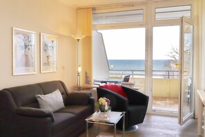 A modern living room with a sofa, armchair, and a small table with a vase. A large window offers a view of the sea.
