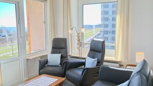Quarto de hotel moderno com duas poltronas reclináveis, uma mesa de madeira com toalha quadriculada e janelas amplas com vista para a paisagem urbana.