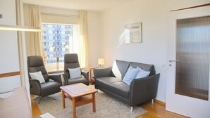 Uma sala de estar moderna com um sofá cinzento, duas poltronas, uma pequena mesa de madeira e uma janela com cortinas.