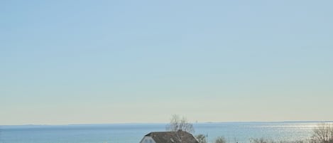A coastal house with a view of the sea, a fenced park area, and a clear sky.