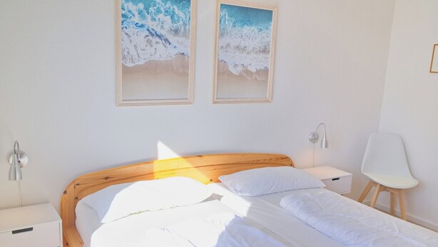 Quarto com cama de madeira, roupa de cama branca, duas molduras na parede, mesa de cabeceira e uma cadeira.