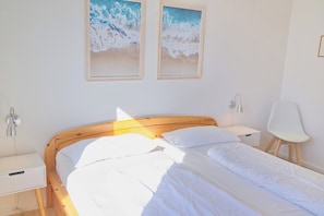 A bedroom with a wooden bed, white bedding, two framed pictures on the wall, a bedside table, and a chair. - Hanseat II, Apartment 148, Wi-Fi included (Grömitz)