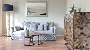 A modern living room with a grey sofa, a round wooden coffee table, a floor lamp, a large mirror, and a perforated wooden partition. - House Baltic App. 40 incl. WLAN (Grömitz)
