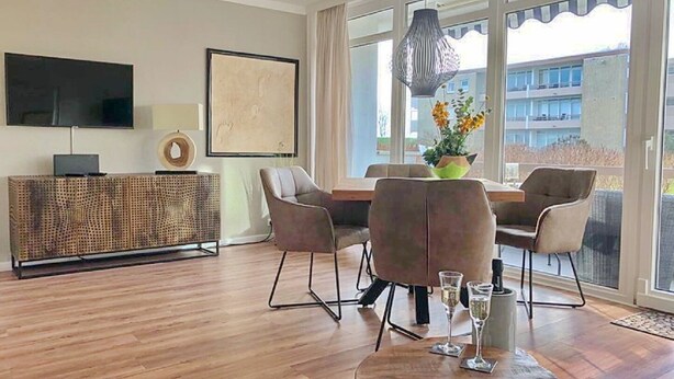 A modern living room with a wooden floor, a flat-screen TV mounted on the wall, a round coffee table with glasses, and a dining area with chairs and a table. - House Baltic App. 40 incl. WLAN (Grömitz)