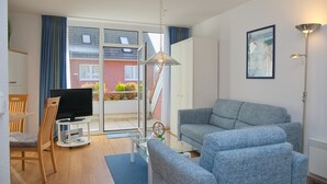 Un salon moderne avec un canapé gris, un téléviseur posé sur un meuble, une table basse en verre et une grande fenêtre donnant sur un bâtiment en briques rouges et sur la verdure environnante.