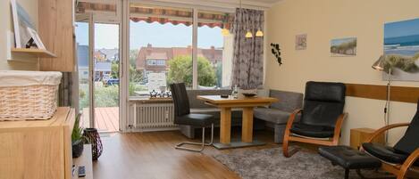 A modern living room with a wooden floor, a dining table, a sofa, and two chairs. There is a window with curtains, a radiator, and a view of the outside.