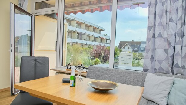 Un salon moderne avec une table en bois, un canapé et une vue sur un immeuble d’habitation, visible à travers de grandes fenêtres.