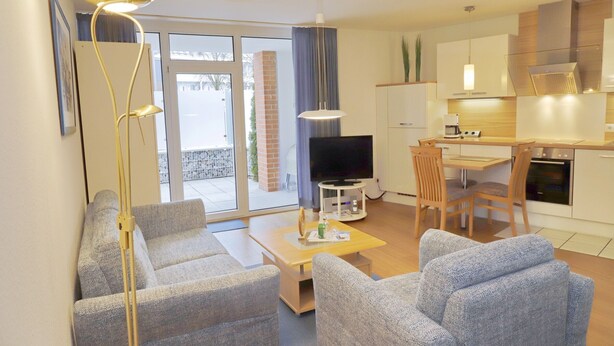 A modern living room with a sofa, a coffee table, and a TV. There's a dining area with a table and chairs. The kitchen is visible with cabinets and a stove. - House Seerose App. 2 incl. WLAN (Grömitz)