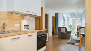 A modern kitchen with white cabinets, a built-in oven, and a coffee maker. There is a dining area with a table and chairs, and a view of the outside through large windows. - House Seerose App. 7 incl. WLAN (Grömitz)
