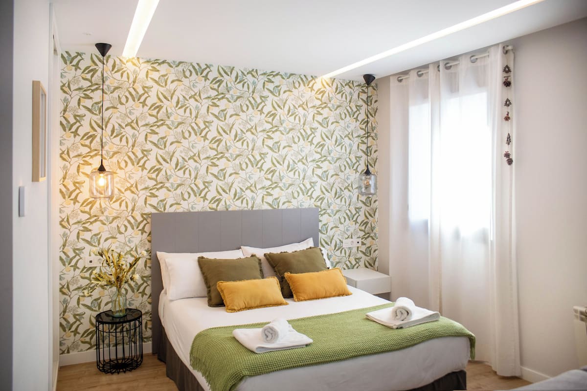 A neatly arranged bedroom with a bed, pillows, a bedside table, and a wall with a leafy pattern.