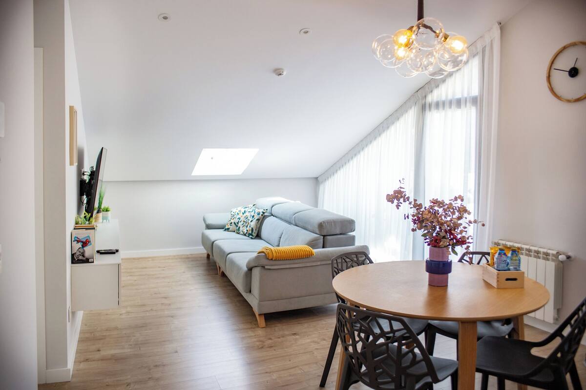 A modern living room with a sofa, a dining table with chairs, a pendant light, and a large window with curtains.
