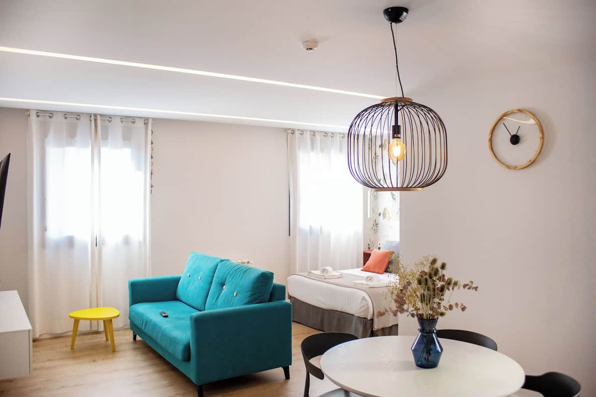 A modern living room with a teal sofa, a small yellow table, a round white table with a vase, and a hanging light fixture.