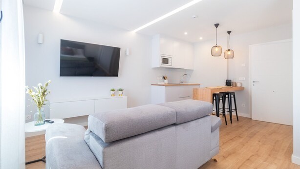 A modern apartment with a grey sofa, a TV mounted on the wall, a kitchen area with a microwave, and a dining table with stools. - A Casa Das Letras (Cangas)