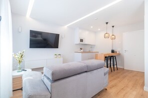 Un apartamento moderno con un sofá gris, un televisor instalado en la pared, una zona de cocina con microondas y una mesa de comedor con taburetes.