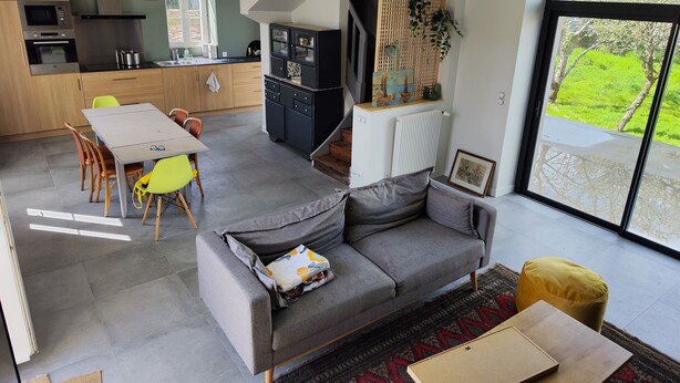 Une cuisine moderne dotée d’appareils électroménagers intégrés, un coin repas avec une table et des chaises, un canapé gris et une grande porte-fenêtre coulissante donnant sur un espace extérieur verdoyant.