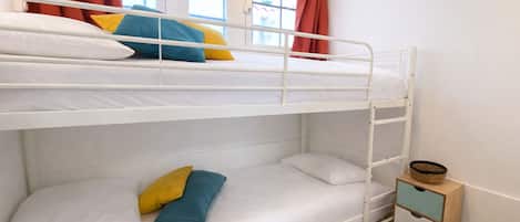 Een slaapkamer met stapelbed, wit beddengoed, een houten nachtkastje en rode gordijnen.