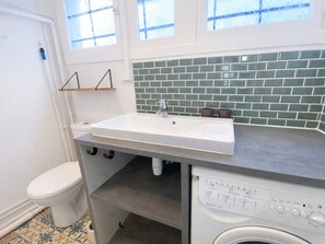 A bathroom with a white sink, a toilet, a washing machine, and a window with a view of the outside.