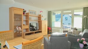 A modern living room with a wooden entertainment unit, a flat-screen TV, wicker furniture, and a view of a building outside. - House Belvedere App. 23 WLAN incl. (Grömitz)