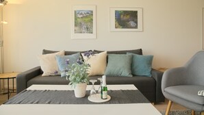 Un salon moderne avec un canapé gris, une plante en pot, une bouteille de vin et deux tableaux représentant des paysages, encadrés et accrochés au mur.