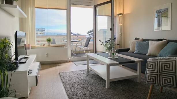 Un salon moderne avec un canapé, une table basse et un téléviseur posé sur un meuble. Il y a un balcon avec vue sur la ville.
