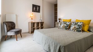Un dormitorio con cama, una silla, mesita de noche, lámpara, estante y un cuadro en la pared.