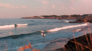 Escena costera con un surfista en la playa, olas y un pueblo lejano con edificios y un faro.