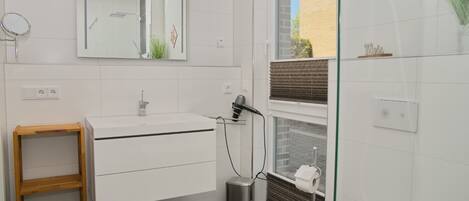 A modern bathroom with a white sink, mirror, and toilet. There is a small trash bin and a wooden shelf.