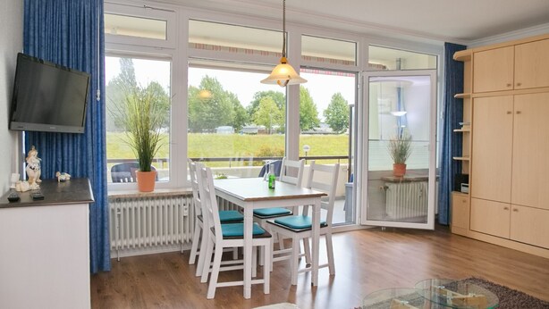 Un espace repas comprenant une table et des chaises, un téléviseur fixé au mur et une vue sur l’extérieur grâce à de grandes fenêtres.