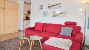 Un salon avec un canapé rouge, une table basse en verre et un plaid blanc. On y trouve des tableaux accrochés au mur et une lampe sur pied.