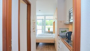 Un couloir étroit mène à une chambre dotée d’une fenêtre, à un coin cuisine équipé d’un four à micro-ondes et à un lit avec un matelas blanc.