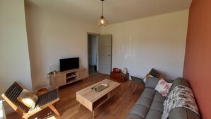 A living room with a wooden coffee table, a sofa, a TV on a wooden stand, and a wooden chair with a woven seat. - Charming T3 in La Roche-Posay Center with Garden, Garage, and Modern Amenities (La Roche-Posay)