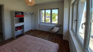 A bedroom with a bed, a wardrobe, a radiator, a chair, and a window with a view of houses. - Charming T3 in La Roche-Posay Center with Garden, Garage, and Modern Amenities (La Roche-Posay)