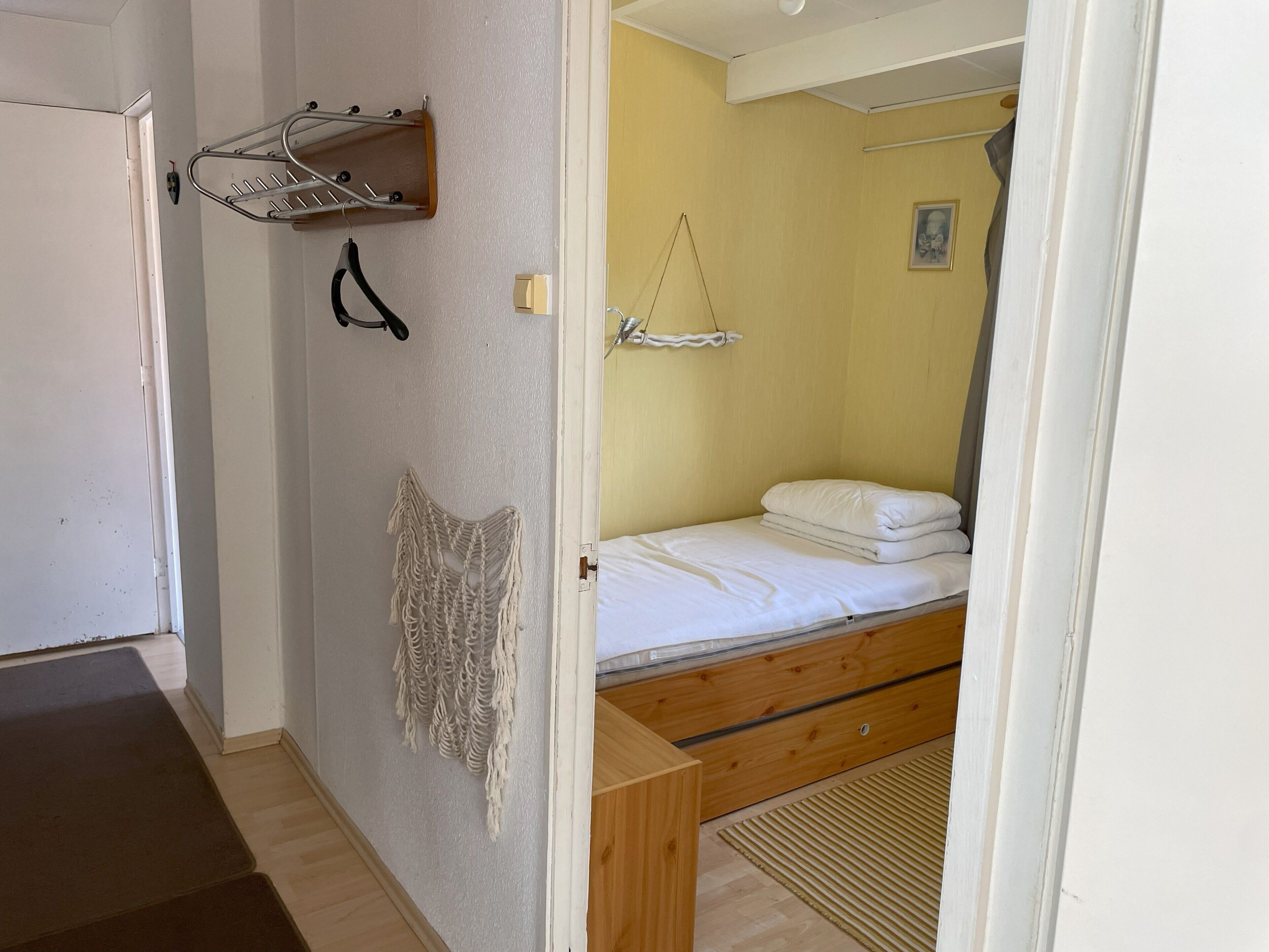 Une petite pièce, de taille modeste, comprenant un lit, une commode en bois, une décoration murale en macramé et un étendoir.
