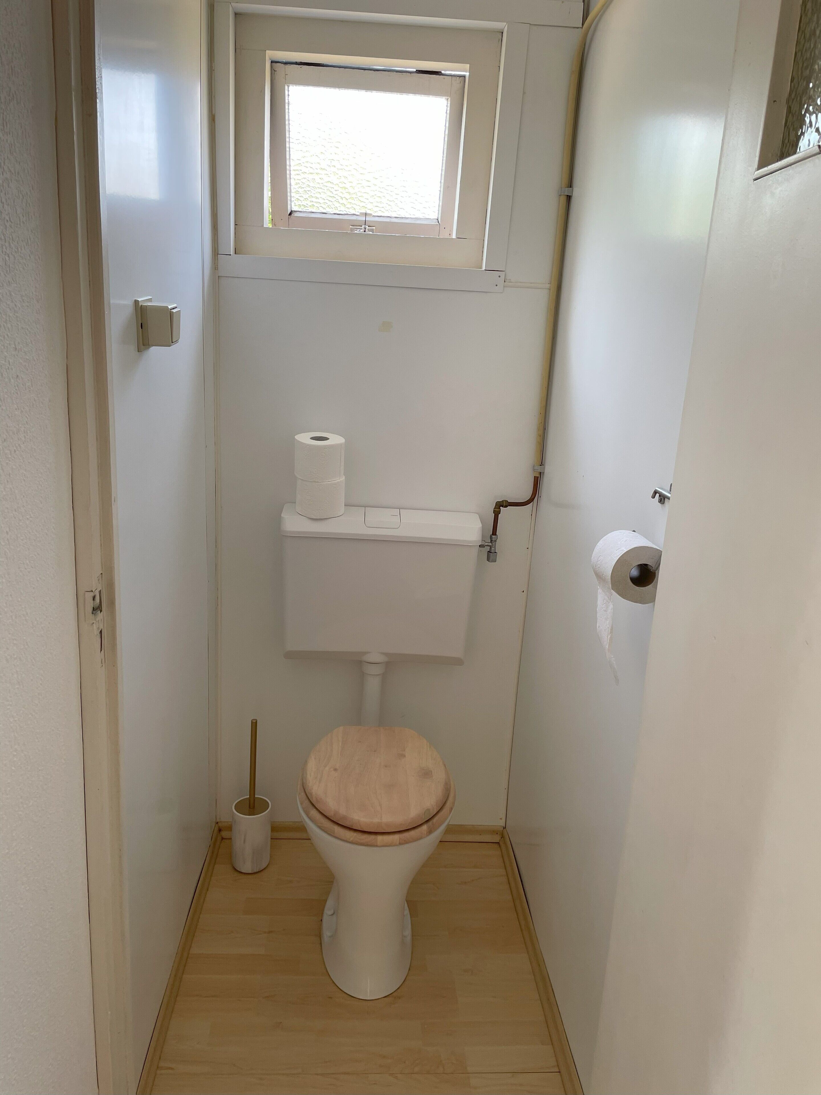 Une petite salle de bain compacte, équipée de toilettes, d’un abattant en bois, d’un rouleau de papier toilette et d’une fenêtre située au-dessus des toilettes.