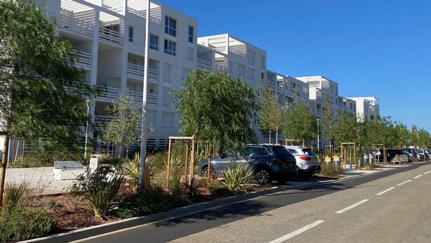 Un immeuble moderne à plusieurs étages, doté de balcons, d’un parking où sont garées des voitures et surplombé d’un ciel bleu clair.