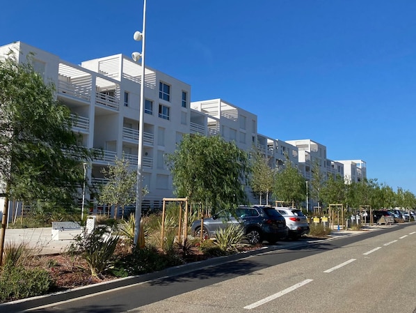 A modern multi-story building with balconies, a parking area with cars, and a clear blue sky.