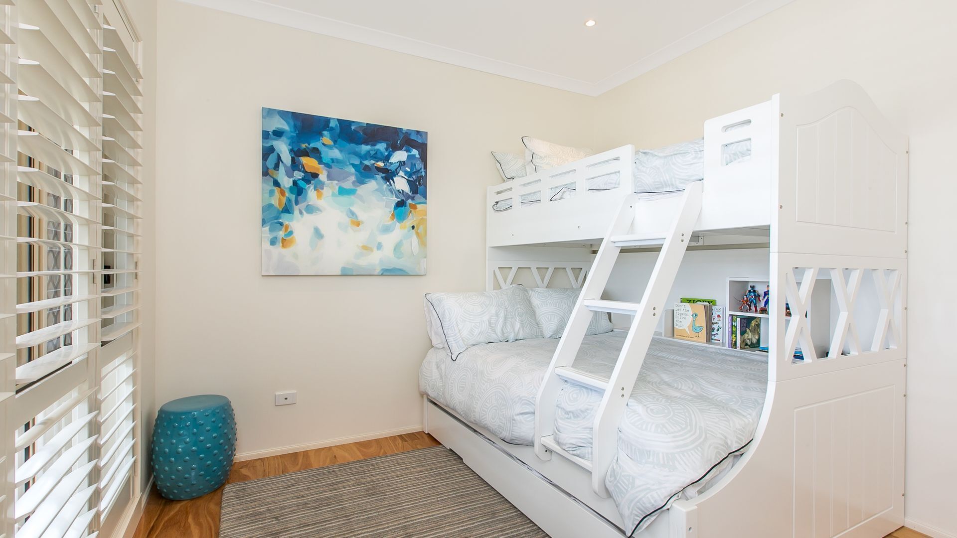Une chambre d’enfant avec des lits superposés, une bibliothèque et un tableau abstrait bleu.