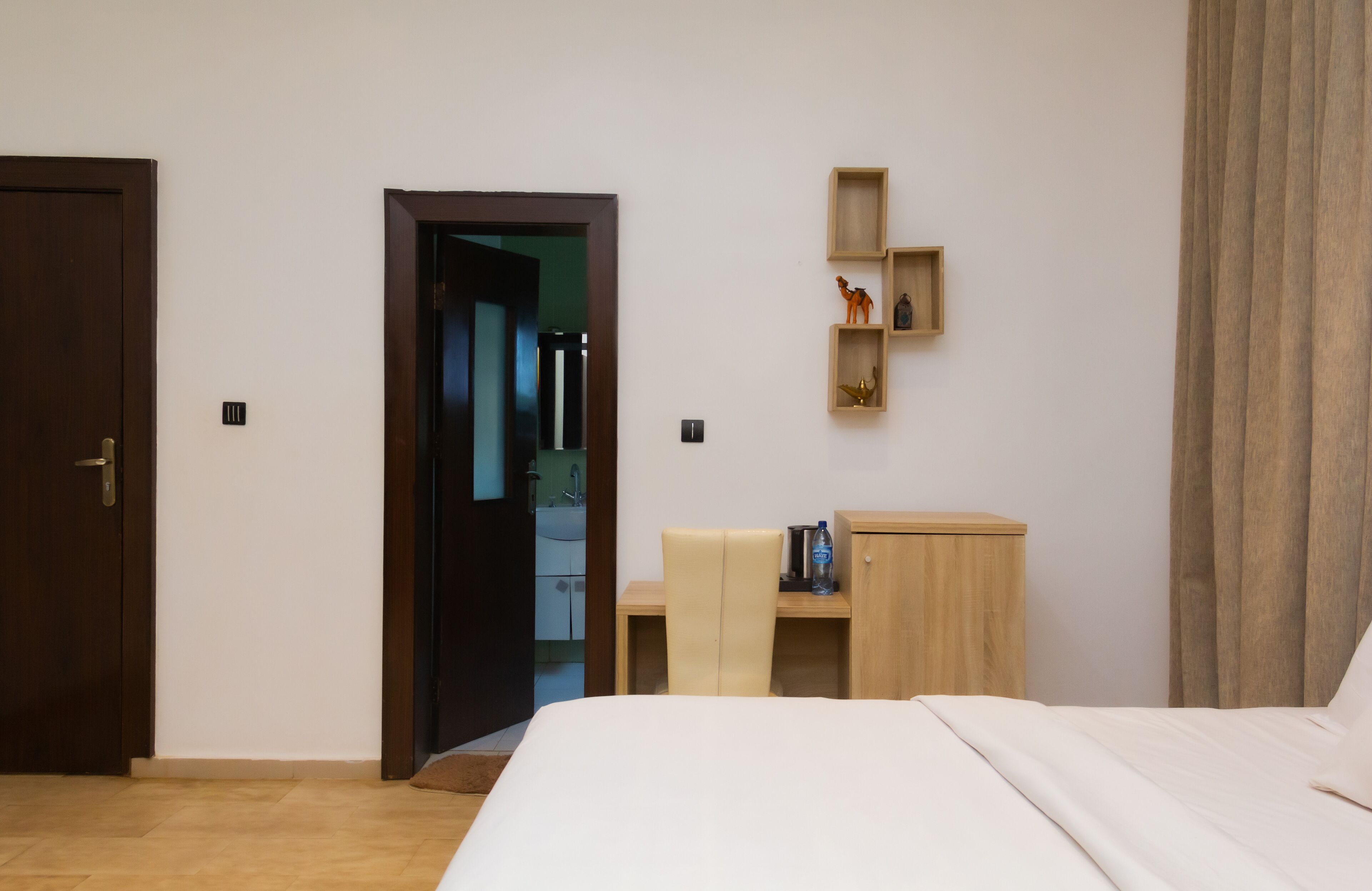 A hotel room with a bed, a desk, a chair, a wardrobe, and a shelf with decorative items.