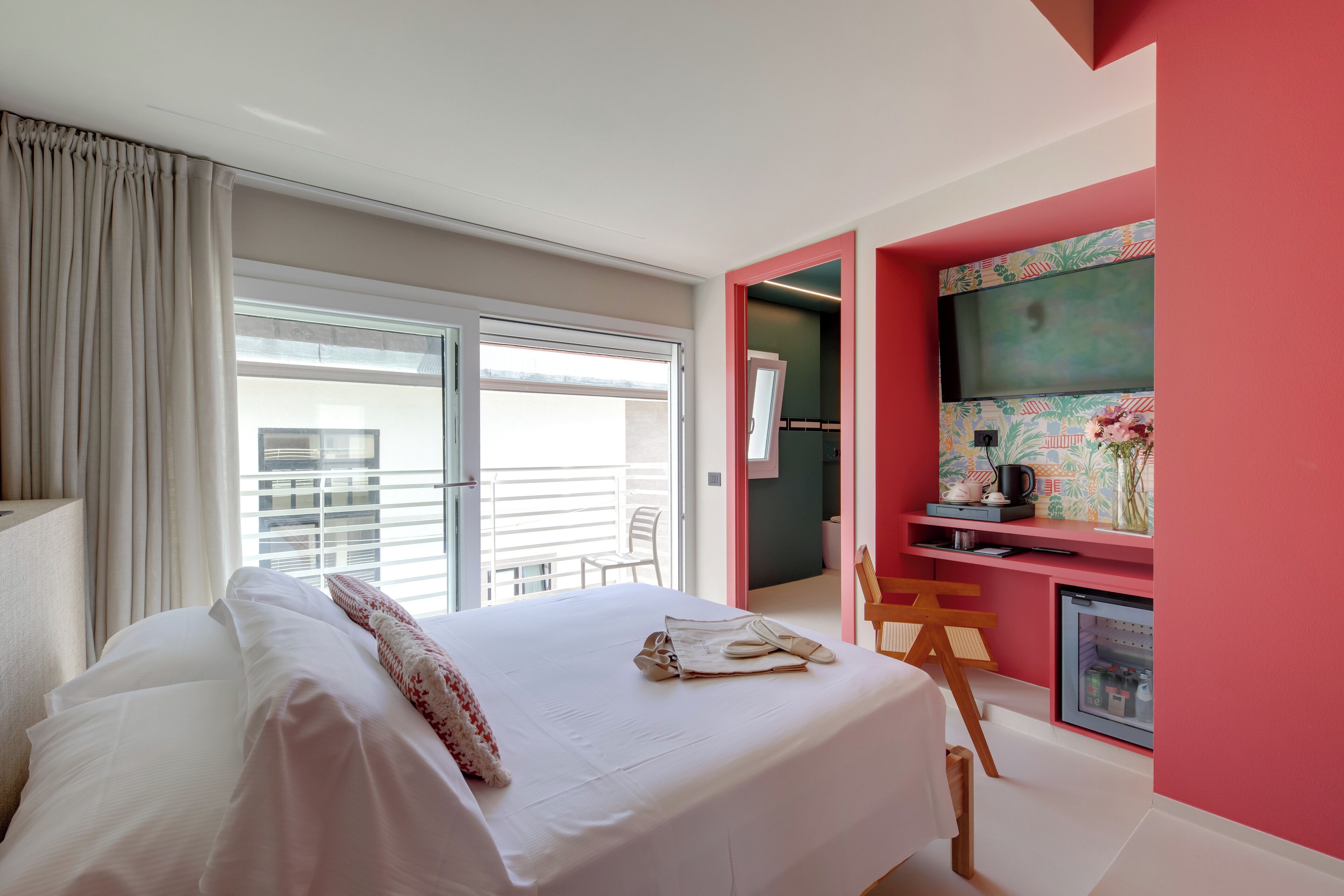 Een hotelkamer met een bed, een bureau, een kleine koelkast en een balkon met uitzicht op de stad.