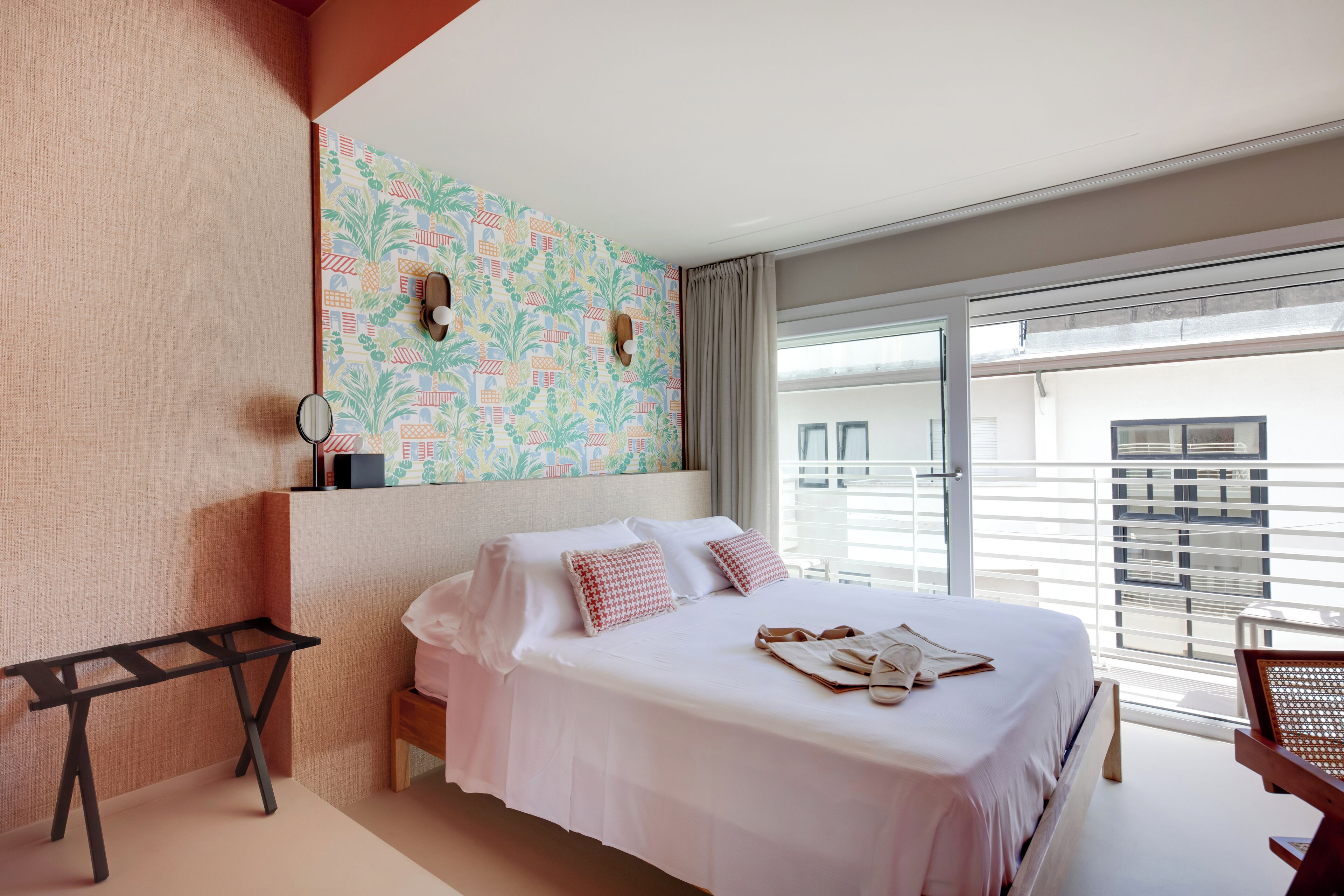 A hotel room with a bed, a small table, a chair, a window with a view of buildings, and a patterned wall.