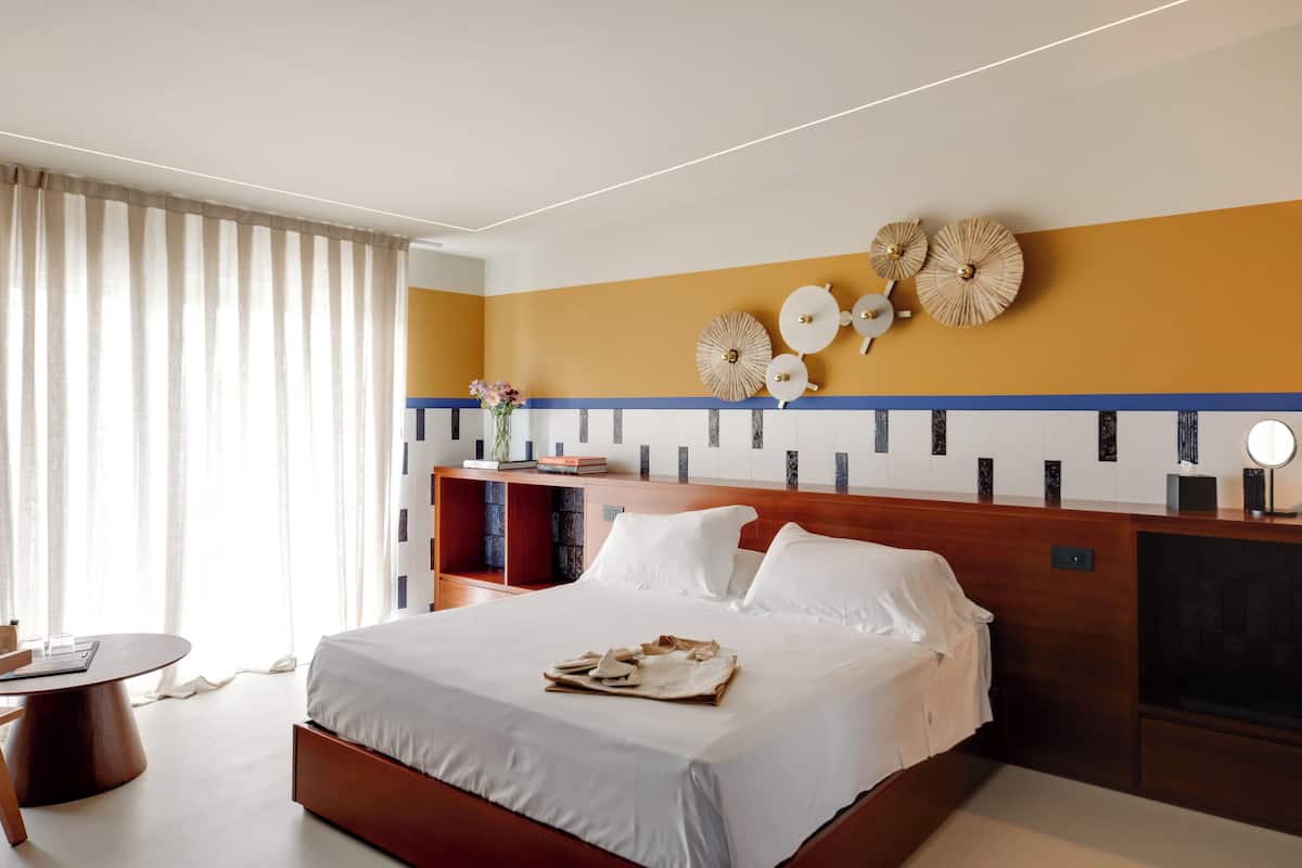 Habitación de hotel con una cama grande, cabecera de madera, mesita de noche con lámpara, una pequeña mesa redonda y una estantería.
