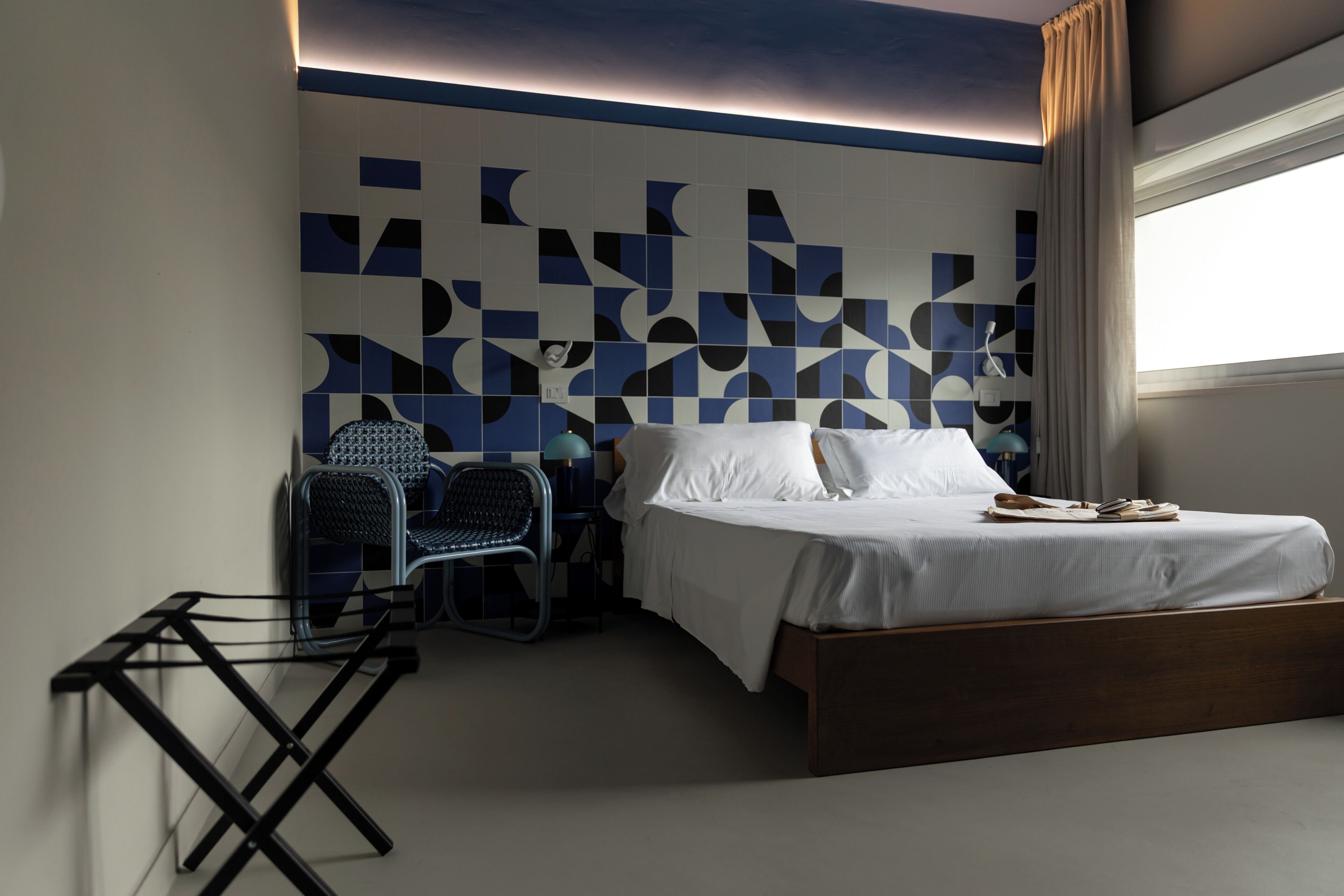 Ein modernes Hotelzimmer mit Wand in geometrischem Muster, einem Einzelbett, einem Sessel und einem kleinen Tisch.