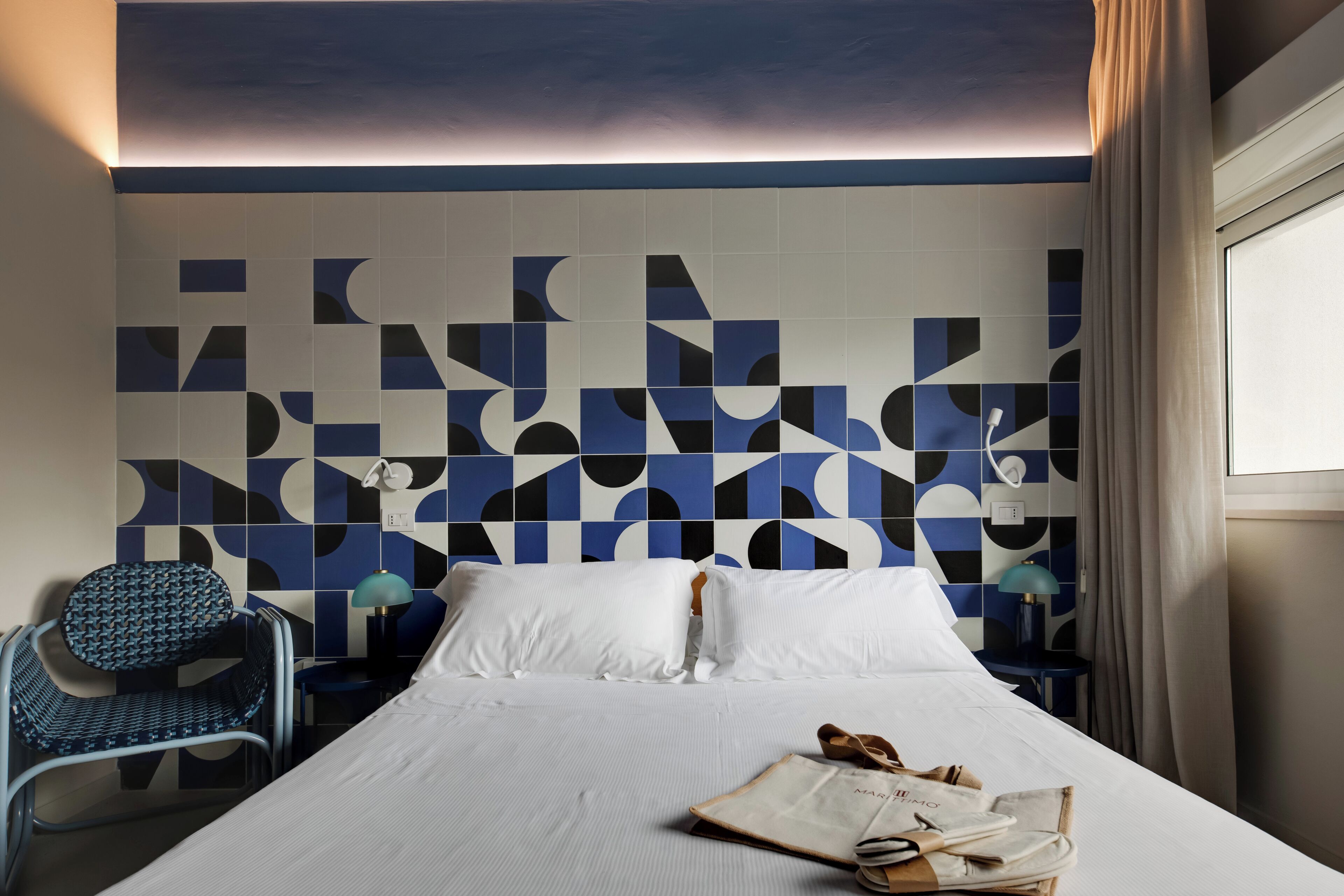 Quarto de hotel com cama, cadeira, mesa de cabeceira e parede com padrões geométricos em azulejos.
