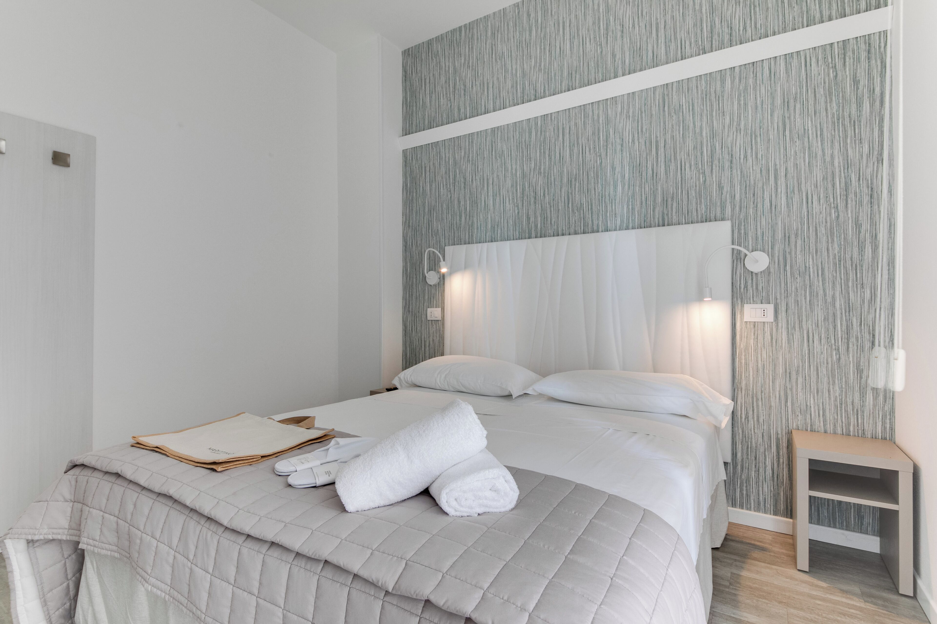 A neatly made bed with white linens and a folded towel, a bedside table with a lamp, and a textured wall behind the bed.