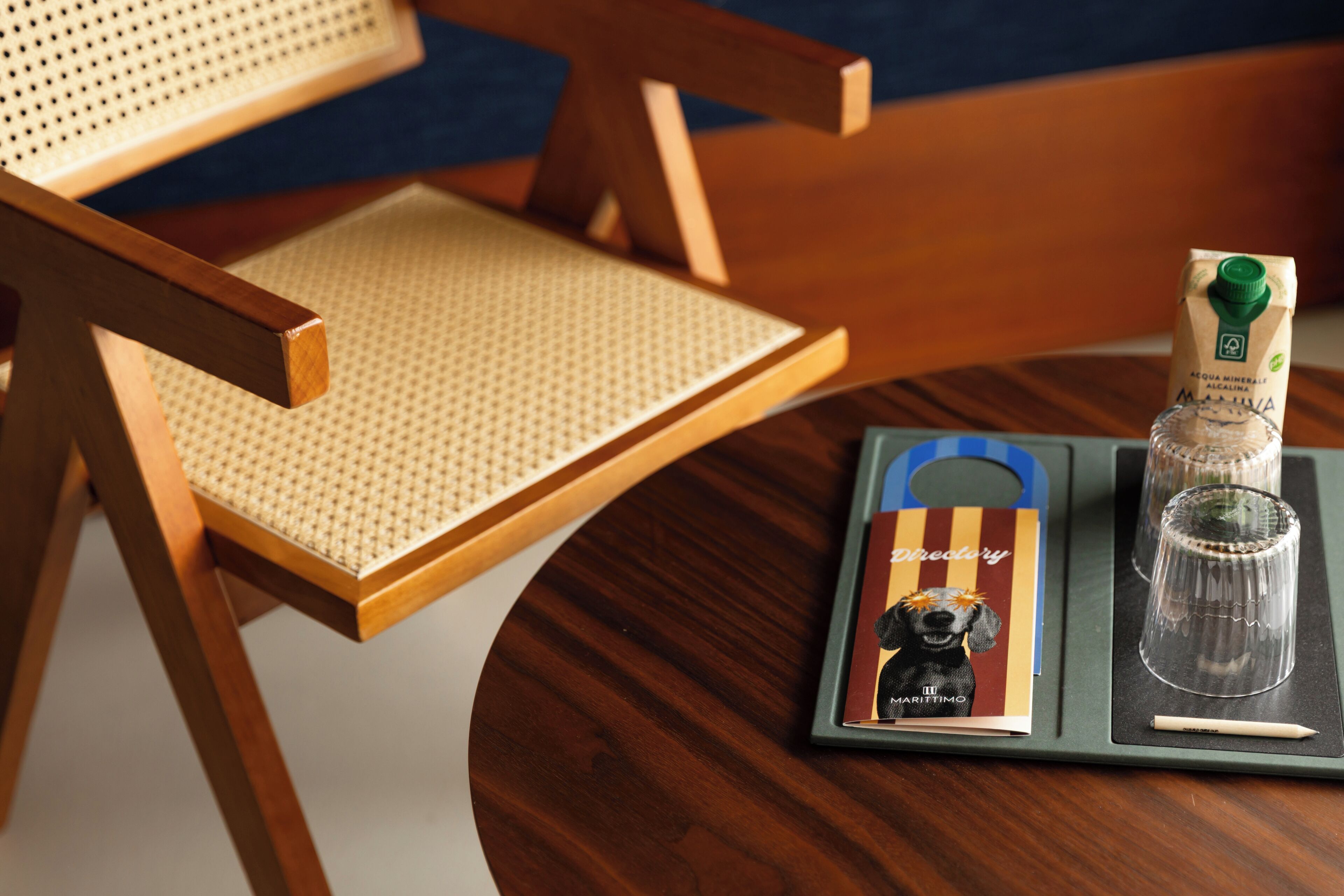 Una silla de madera con asiento tejido, una mesita con una bandeja que sostiene una botella de agua mineral, un vaso y un menú.