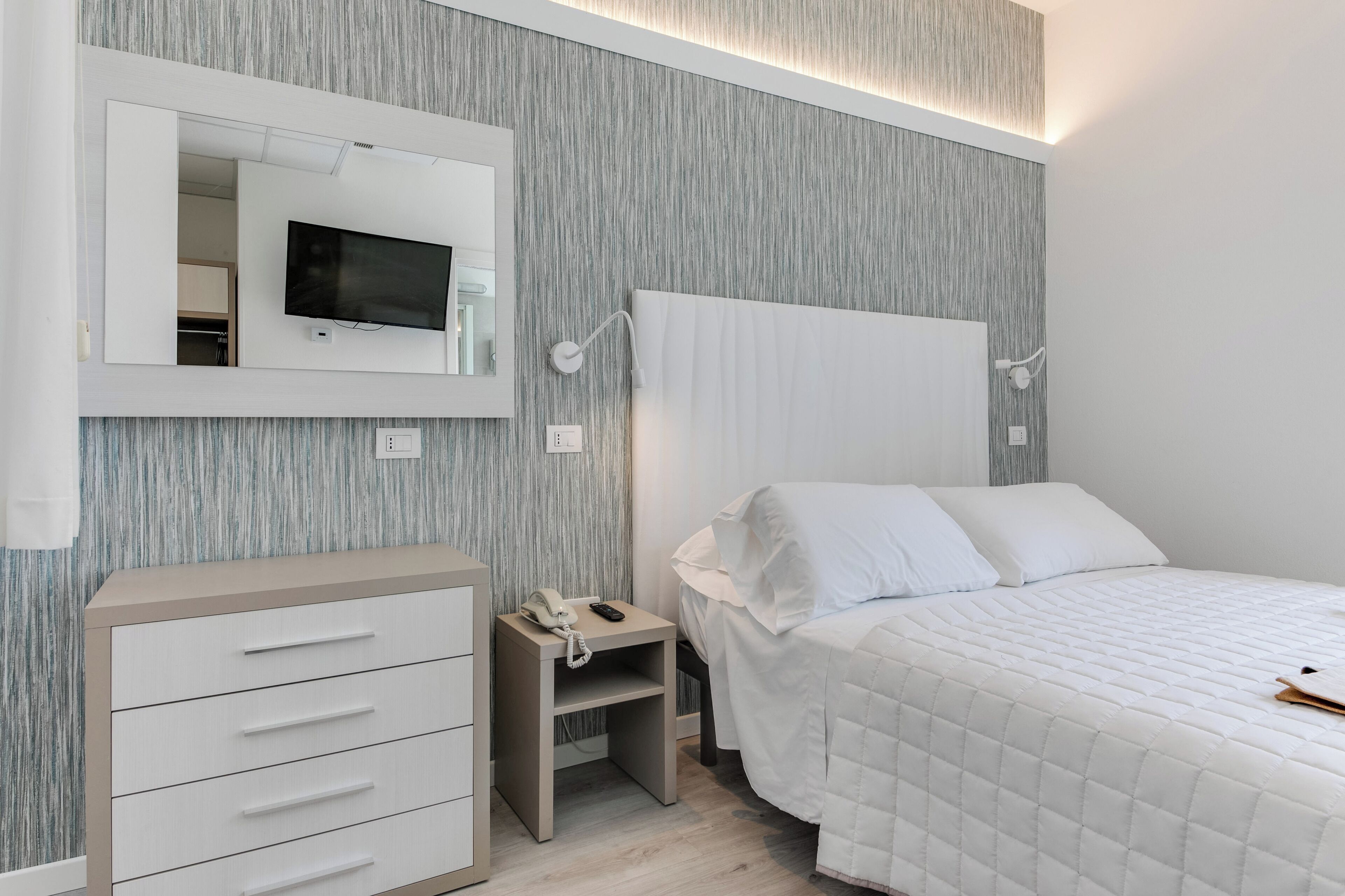 Una camera da letto con un letto, un comodino con un telefono, uno specchio e una TV montata a parete.